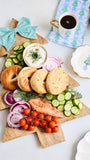 A Christmas tree-shaped wooden serving board styled with bagel halves, cucumber slices, red onion, dill, a small bowl of cream cheese, sliced salmon, cherry tomatoes, and a striped teal bow, arranged on a white tabletop with a cup of coffee and decorative plates nearby.