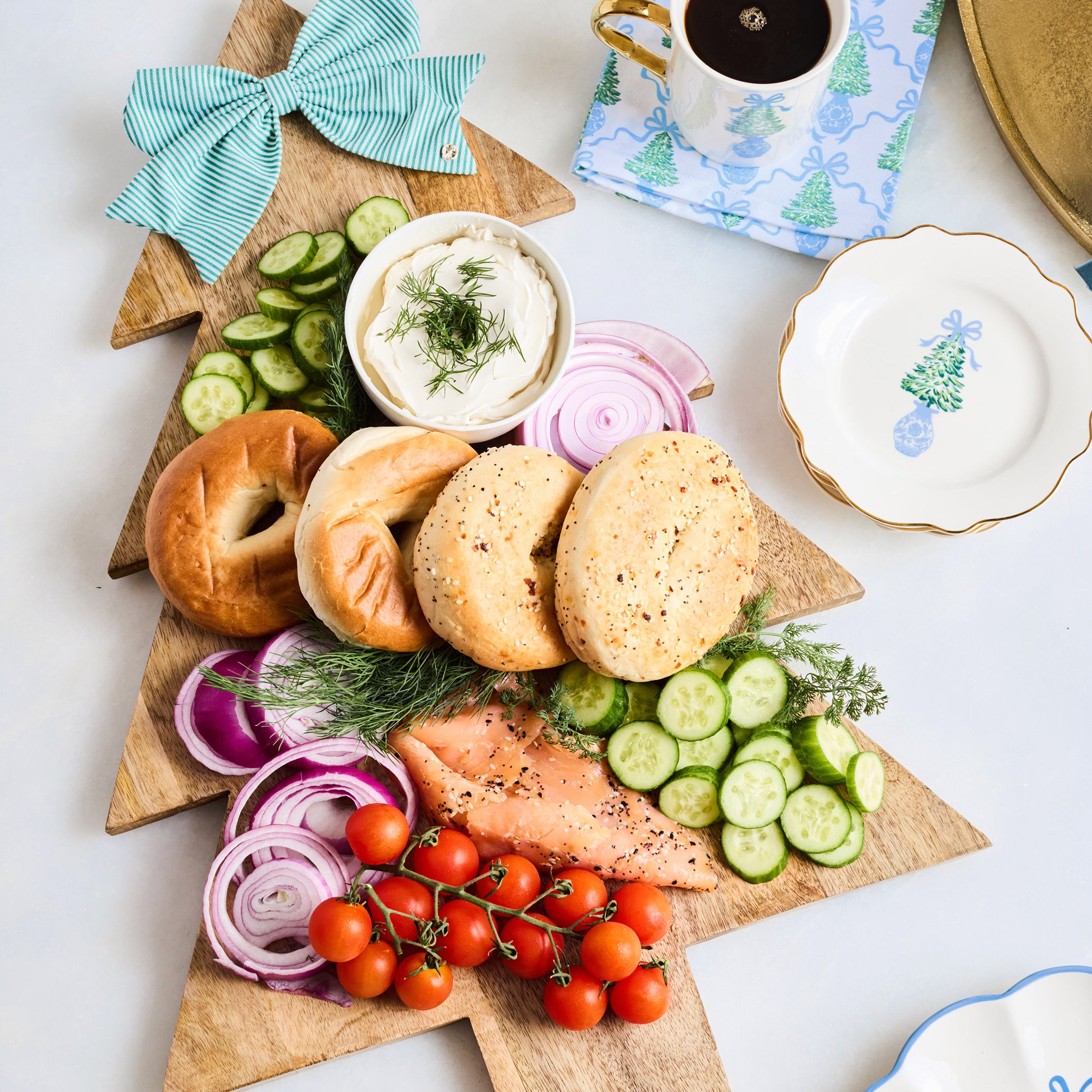 A Christmas tree-shaped wooden serving board styled with bagel halves, cucumber slices, red onion, dill, a small bowl of cream cheese, sliced salmon, cherry tomatoes, and a striped teal bow, arranged on a white tabletop with a cup of coffee and decorative plates nearby.