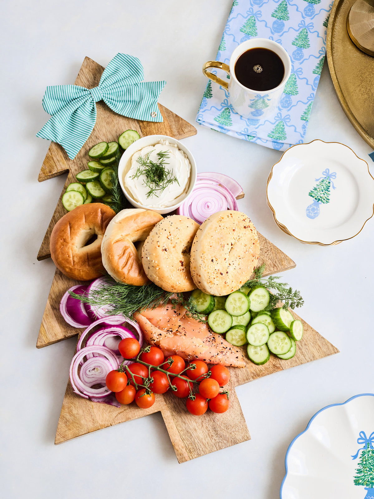 A Christmas tree-shaped wooden serving board styled with bagel halves, cucumber slices, red onion, dill, a small bowl of cream cheese, sliced salmon, cherry tomatoes, and a striped teal bow, arranged on a white tabletop with a cup of coffee and decorative plates nearby.