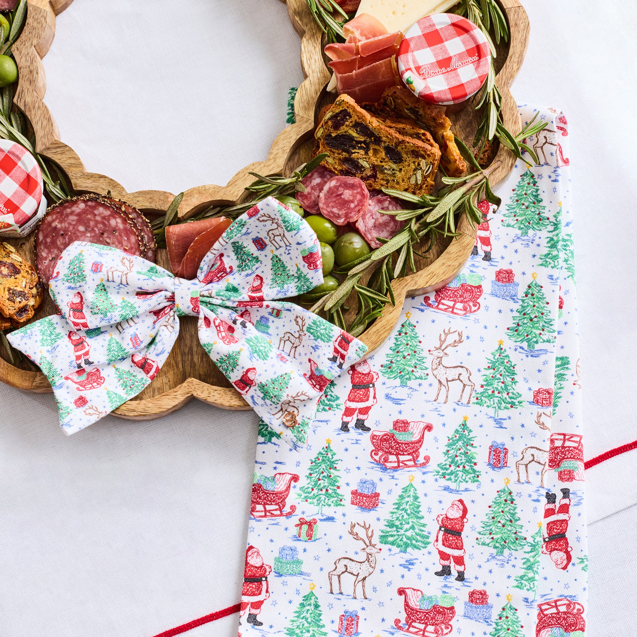 A festive Christmas-themed food charcuterie board arranged on a scalloped wooden tray, with a matching printed bow and folded napkin beside it. The board is topped with salami, prosciutto, cheese, olives, crackers, and greenery, set on a white surface.