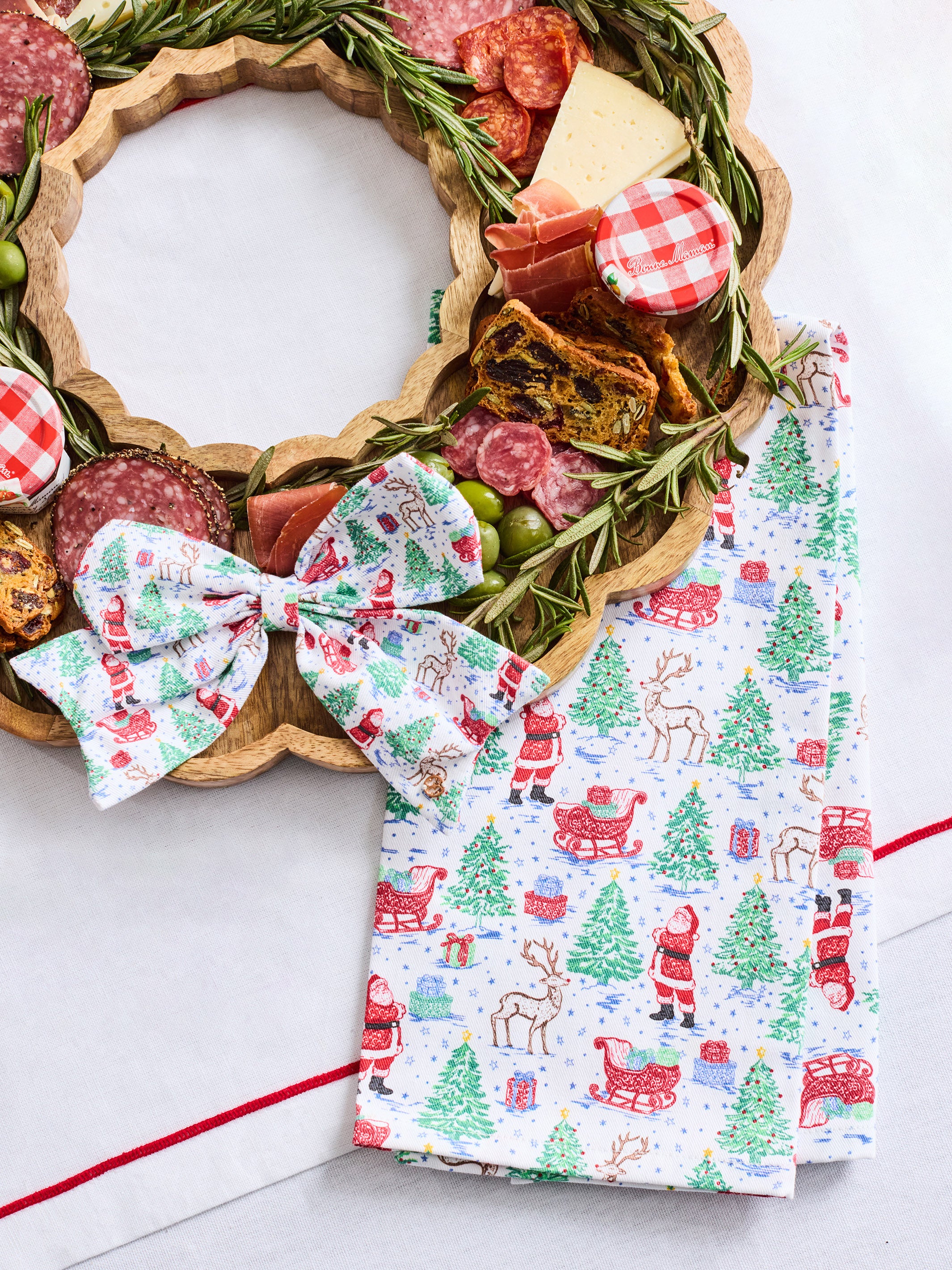 A festive Christmas-themed food charcuterie board arranged on a scalloped wooden tray, with a matching printed bow and folded napkin beside it. The board is topped with salami, prosciutto, cheese, olives, crackers, and greenery, set on a white surface.
