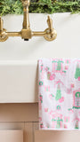 A white waffle-weave kitchen towel with a pink and green Christmas village pattern hangs over the edge of a white sink beneath a brass faucet.