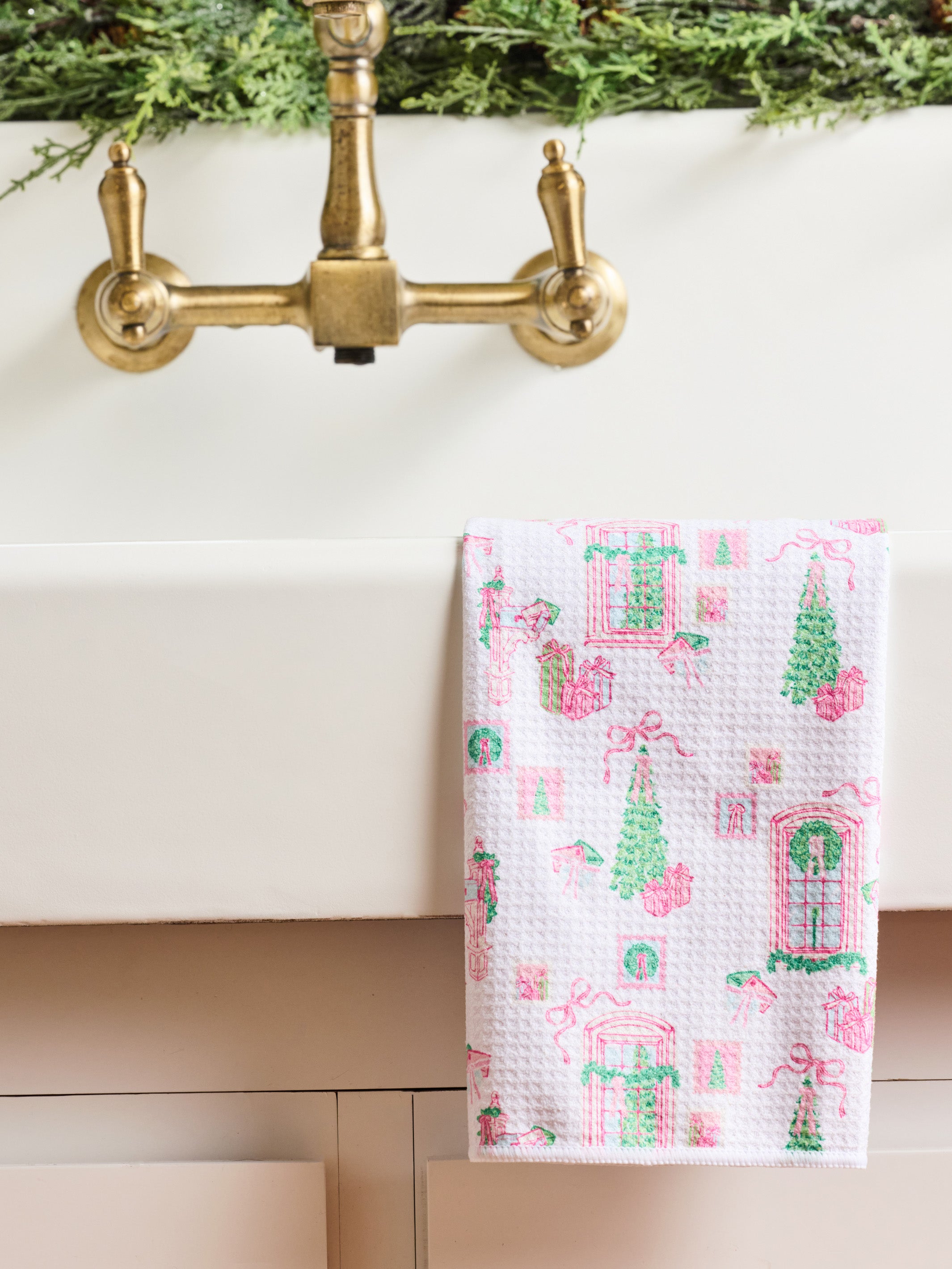 A white waffle-weave kitchen towel with a pink and green Christmas village pattern hangs over the edge of a white sink beneath a brass faucet.