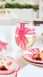 A clear stemless wine glass decorated with a pink and red striped bow design, filled with white wine, set on a white table with holiday-themed plates and cookies.