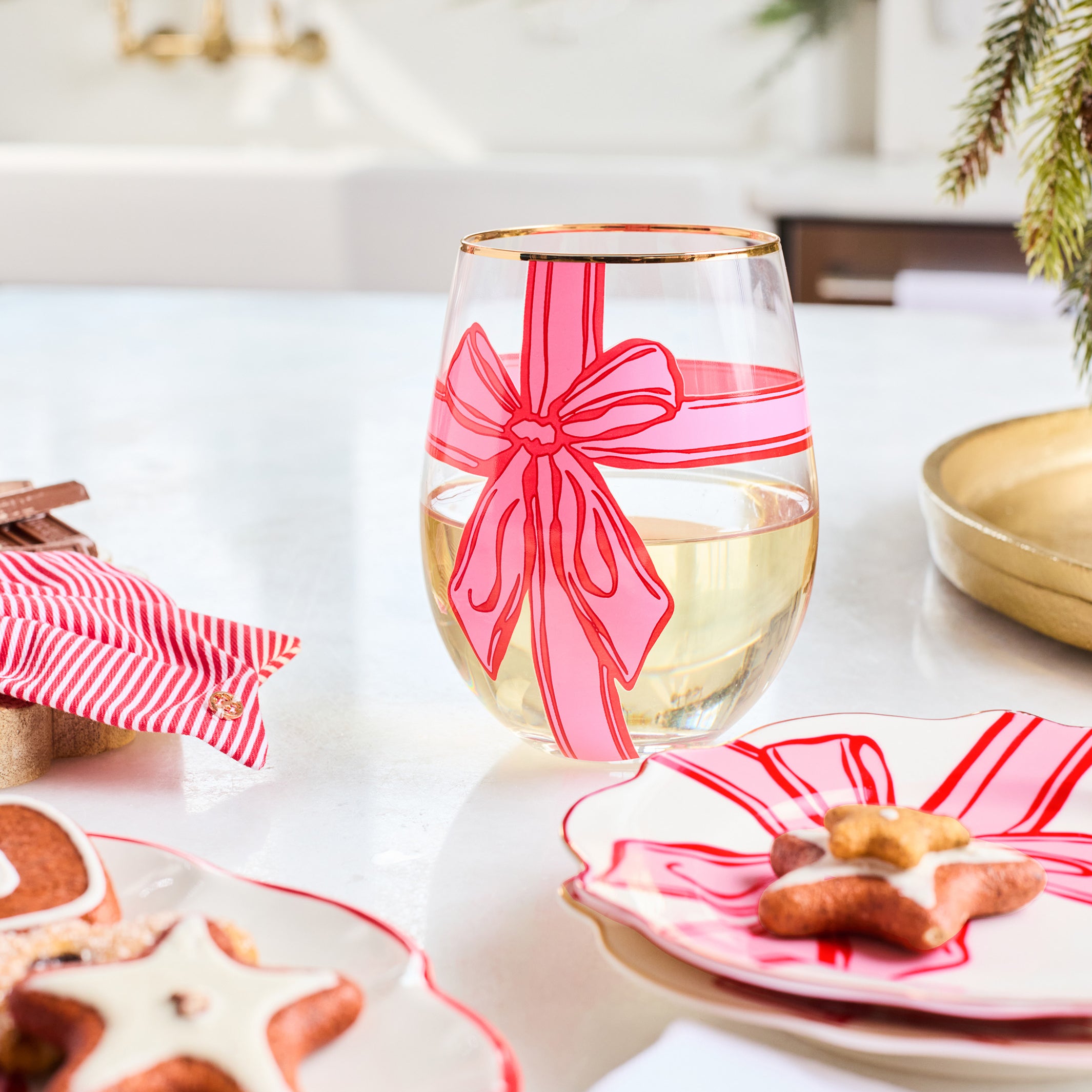 A clear stemless wine glass decorated with a pink and red striped bow design, filled with white wine, set on a white table with holiday-themed plates and cookies.