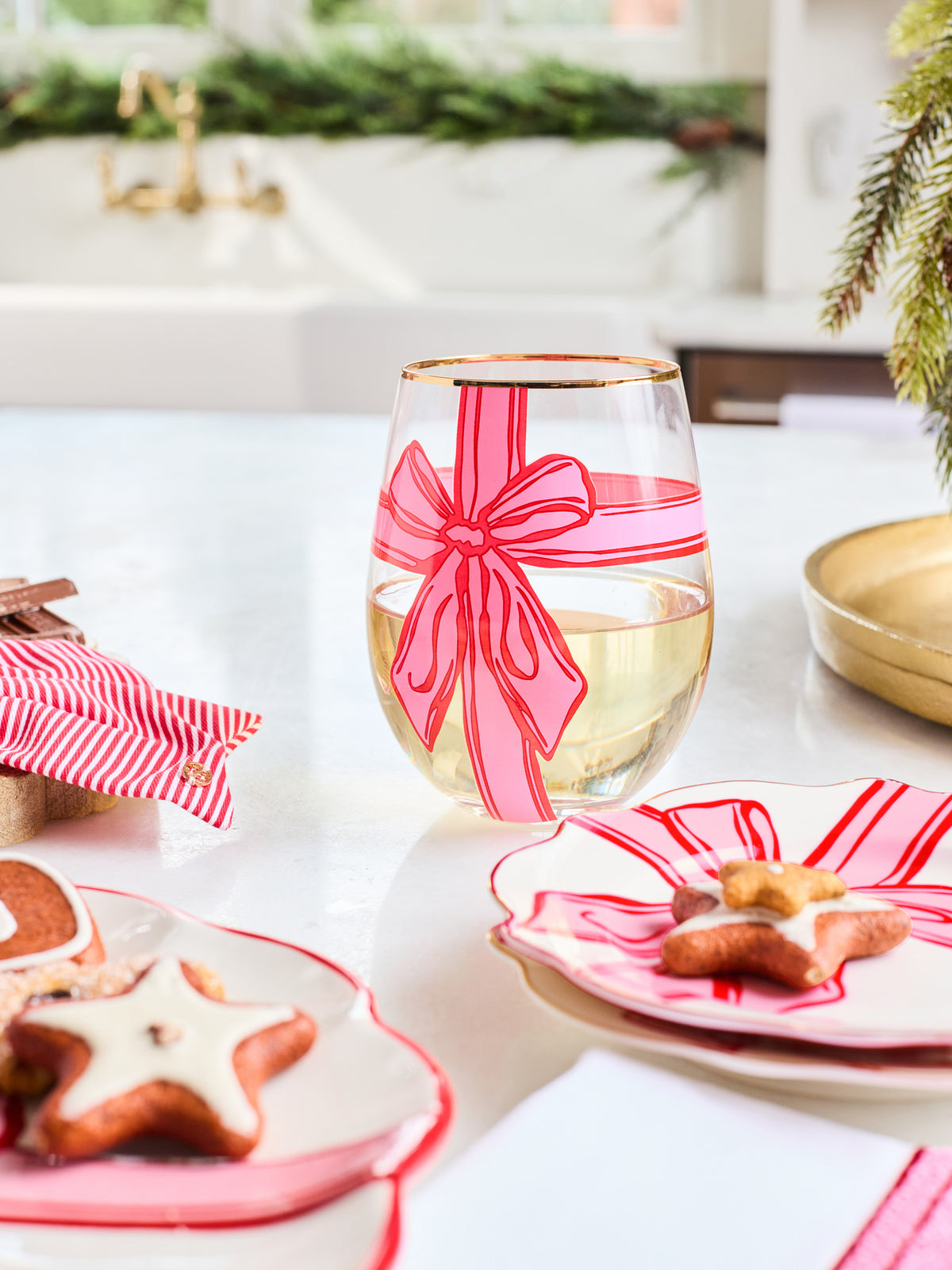 A clear stemless wine glass decorated with a pink and red striped bow design, filled with white wine, set on a white table with holiday-themed plates and cookies.