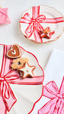 A styled flat-lay of three white ceramic plates with scalloped edges, each decorated with a red and pink gift ribbon pattern. The plates are arranged on a white surface with decorated gingerbread cookies (heart and star shapes) and a striped napkin in the background.