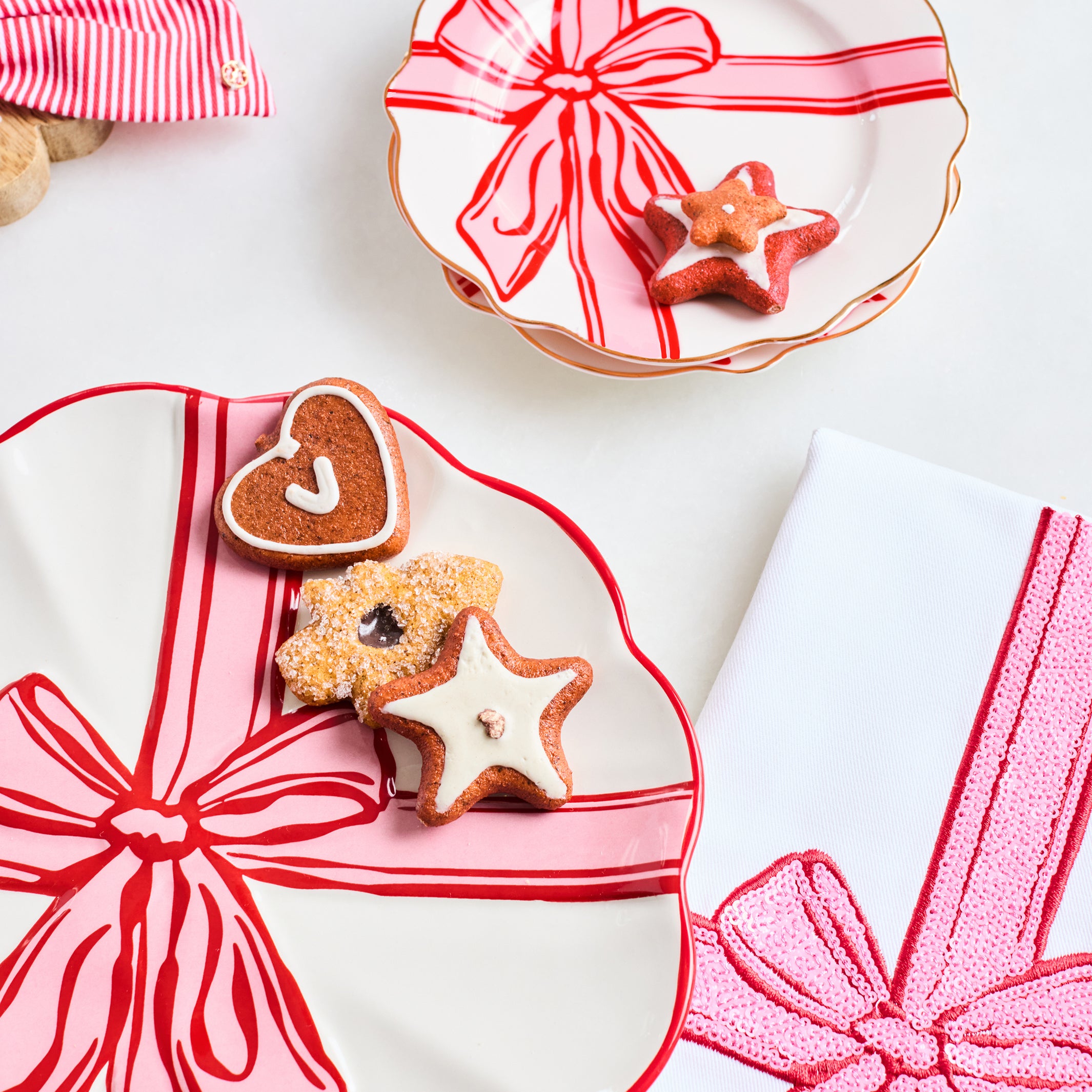 A styled flat-lay of three white ceramic plates with scalloped edges, each decorated with a red and pink gift ribbon pattern. The plates are arranged on a white surface with decorated gingerbread cookies (heart and star shapes) and a striped napkin in the background.