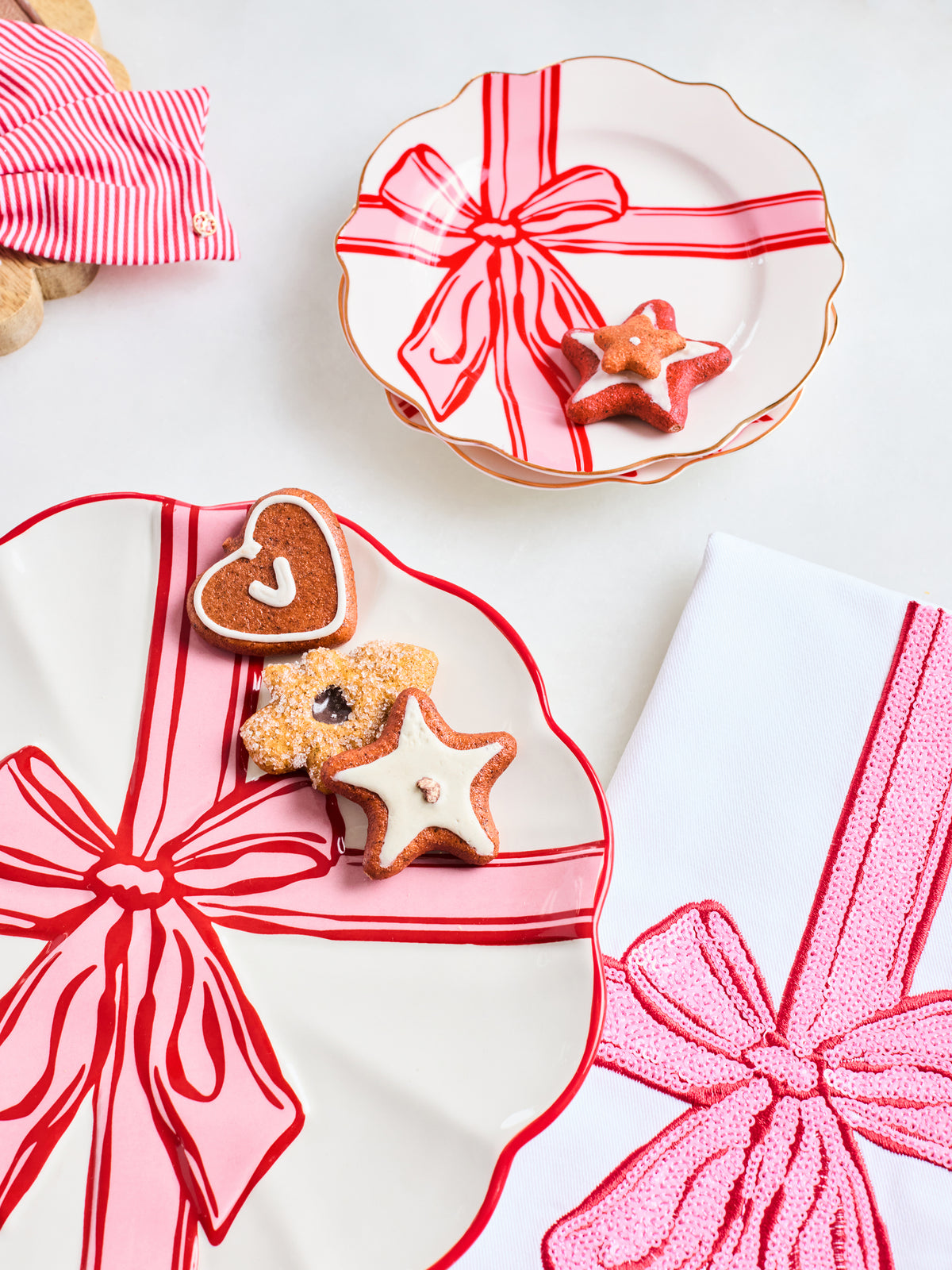A styled flat-lay of three white ceramic plates with scalloped edges, each decorated with a red and pink gift ribbon pattern. The plates are arranged on a white surface with decorated gingerbread cookies (heart and star shapes) and a striped napkin in the background.