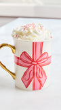 A white ceramic mug with a gold handle, decorated with a pink gift bow graphic, filled with whipped cream and candy canes, sitting on a white surface.