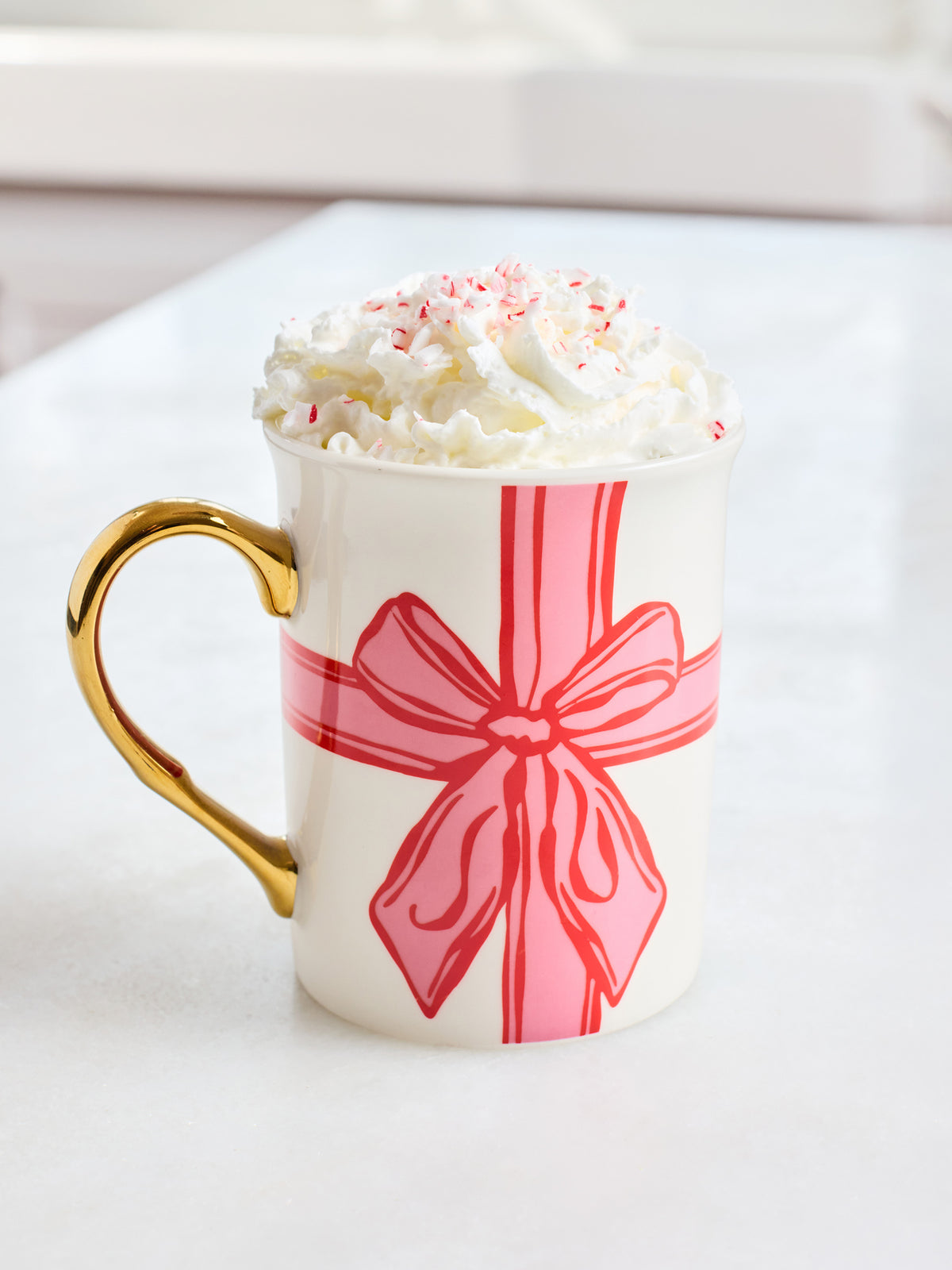 A white ceramic mug with a gold handle, decorated with a pink gift bow graphic, filled with whipped cream and candy canes, sitting on a white surface.