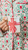 A person wearing a festive Christmas-themed pajama set with a red, green, and pink floral pattern, holding a white mug decorated with a red gift box illustration.