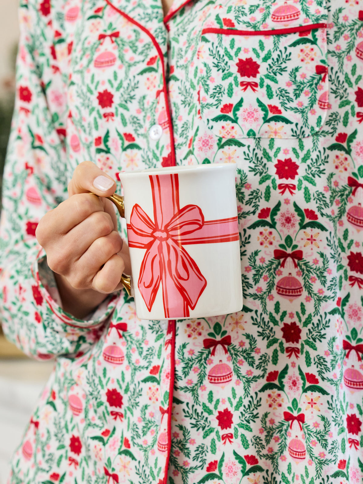 A person wearing a festive Christmas-themed pajama set with a red, green, and pink floral pattern, holding a white mug decorated with a red gift box illustration.