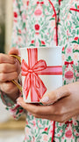 A person in festive floral pajamas holds a white ceramic mug with a gold handle decorated with a red and pink gift box bow design.