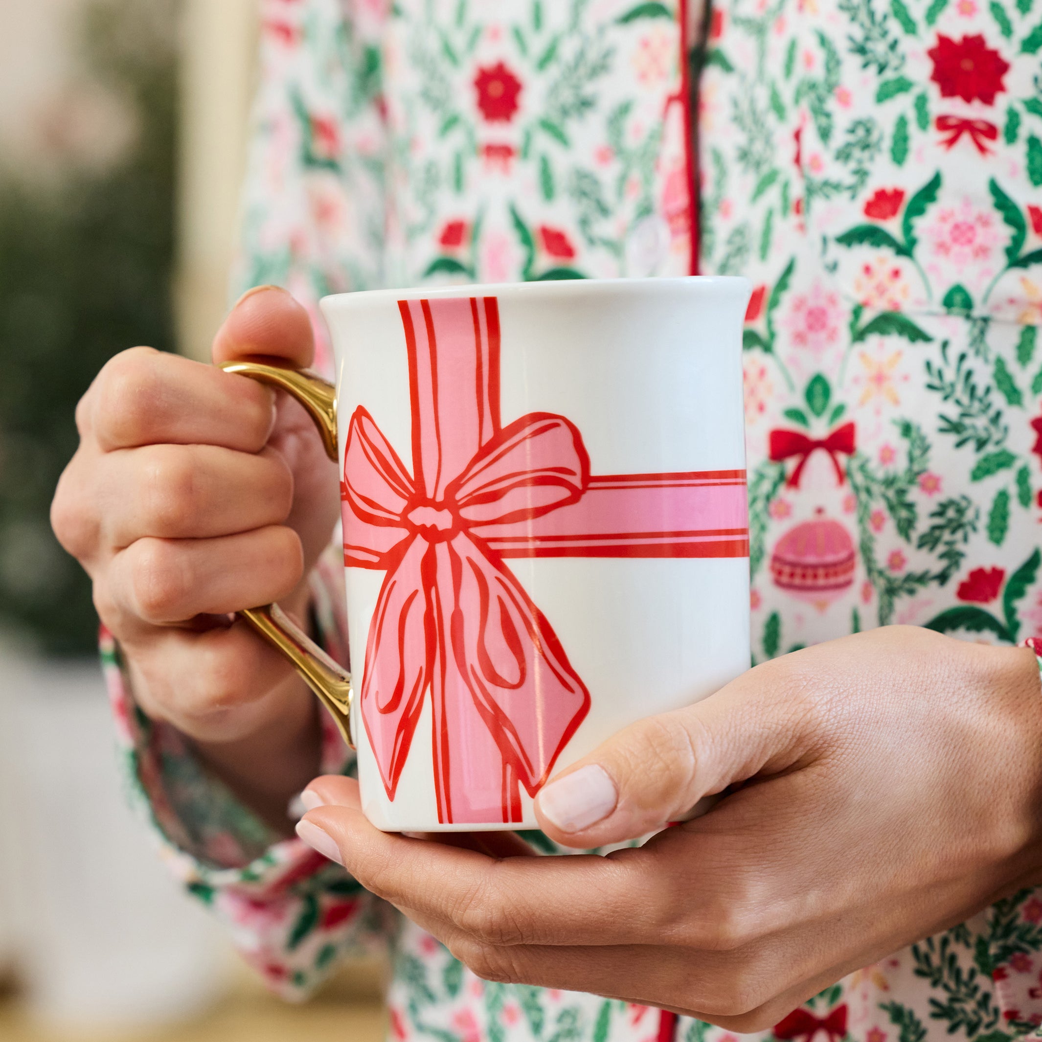 A person in festive floral pajamas holds a white ceramic mug with a gold handle decorated with a red and pink gift box bow design.