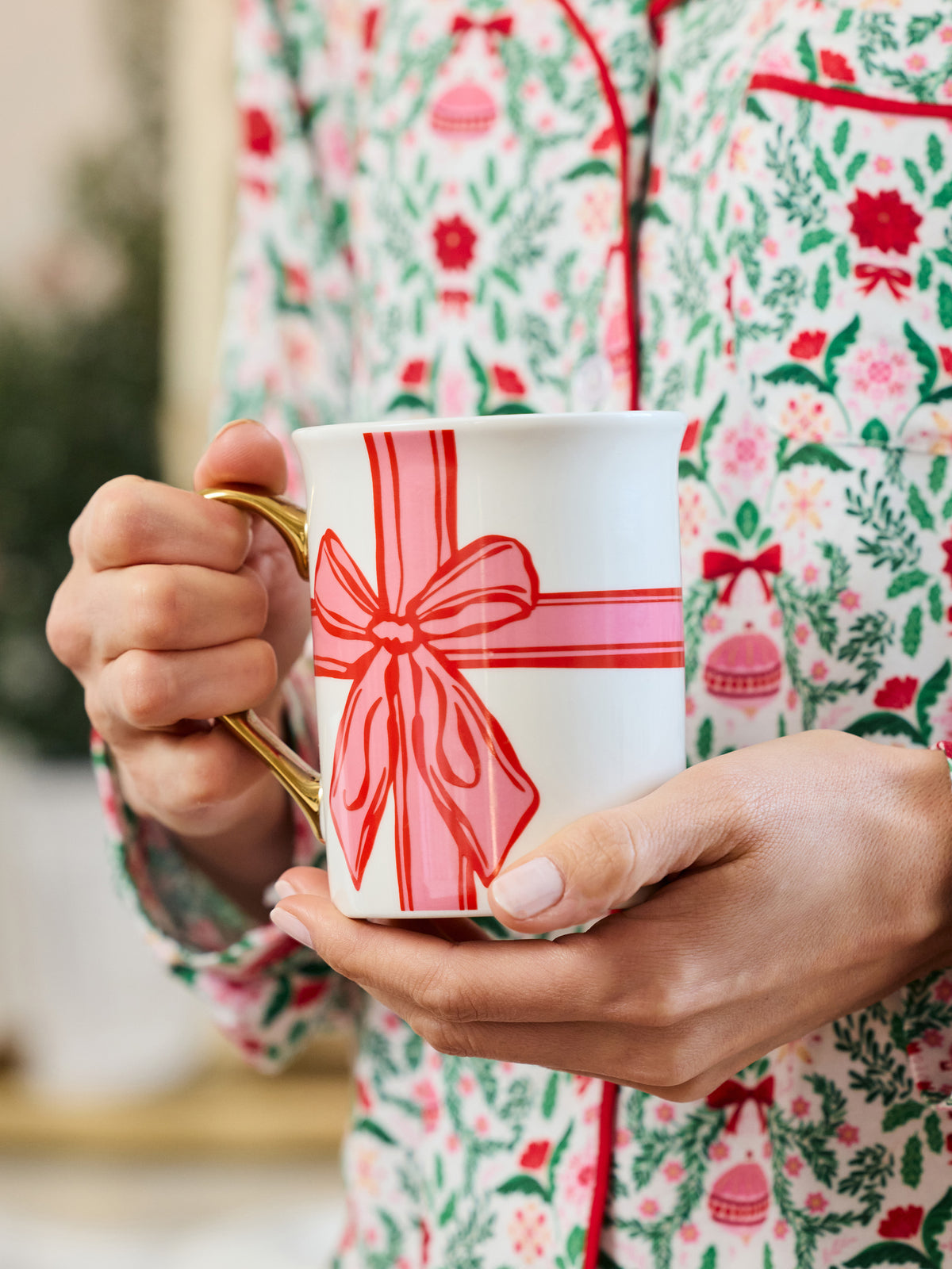 A person in festive floral pajamas holds a white ceramic mug with a gold handle decorated with a red and pink gift box bow design.