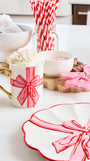 A festive white ceramic mug decorated with a red and pink striped bow and handle, filled with whipped cream and sprinkles, styled on a white countertop with holiday-themed plates, straws, and candy in a bright kitchen setting.