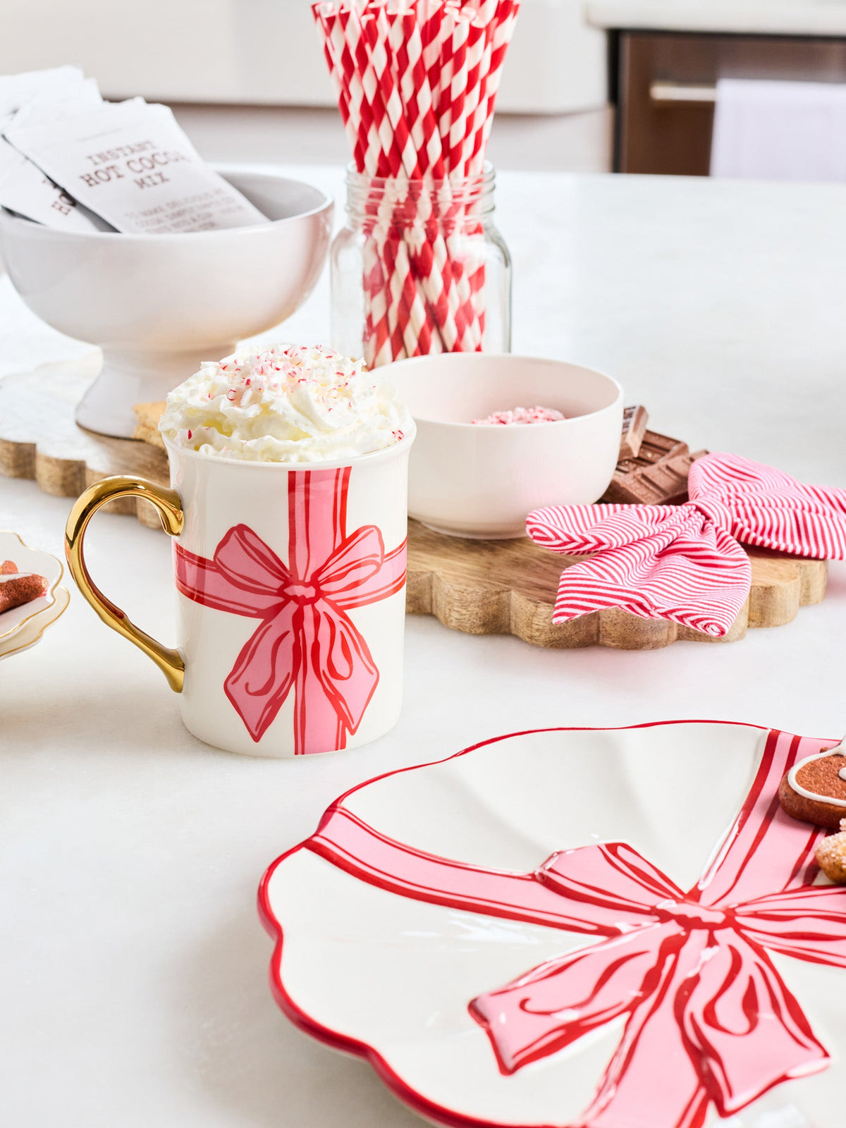 A festive white ceramic mug decorated with a red and pink striped bow and handle, filled with whipped cream and sprinkles, styled on a white countertop with holiday-themed plates, straws, and candy in a bright kitchen setting.