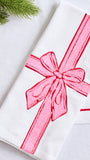 A folded white fabric napkin or towel featuring a large pink embroidered ribbon and bow motif, styled on a light surface with a small green pine branch in the corner.