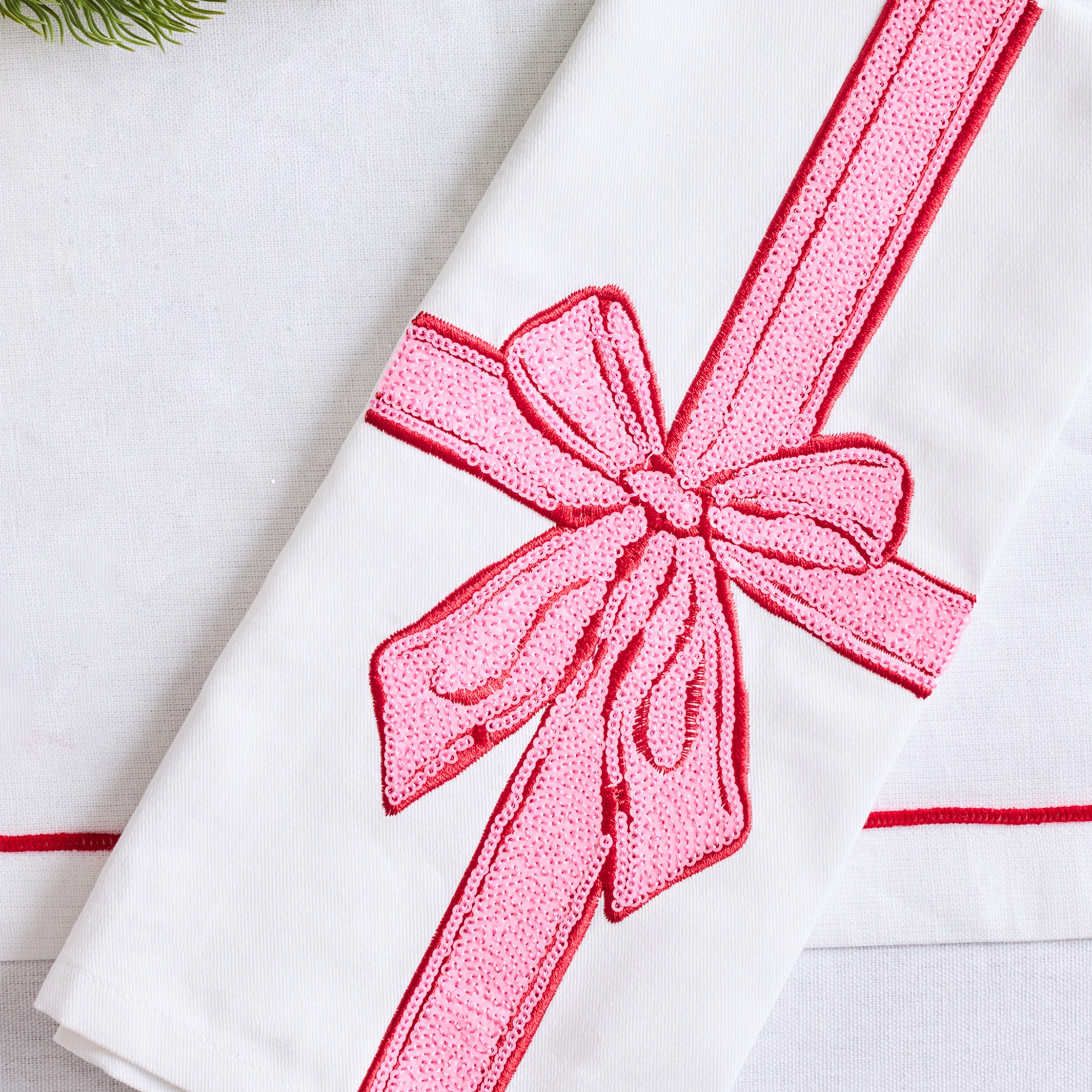 A folded white fabric napkin or towel featuring a large pink embroidered ribbon and bow motif, styled on a light surface with a small green pine branch in the corner.