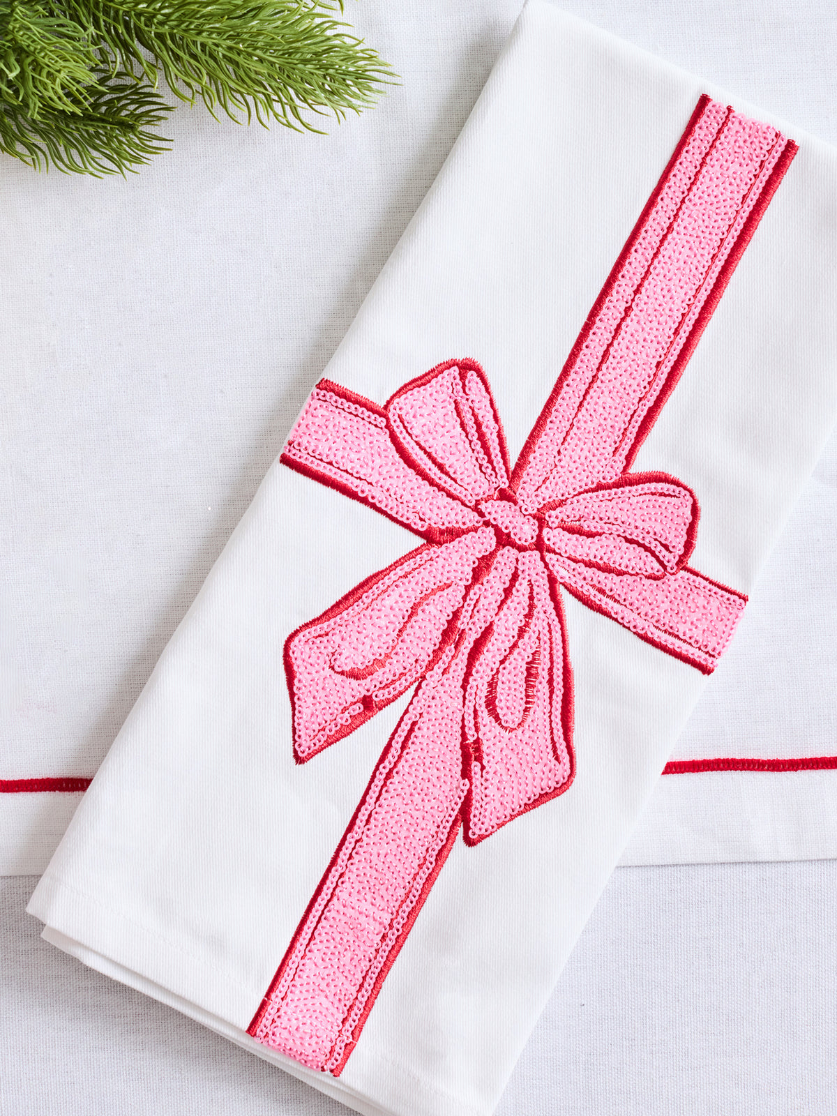 A folded white fabric napkin or towel featuring a large pink embroidered ribbon and bow motif, styled on a light surface with a small green pine branch in the corner.