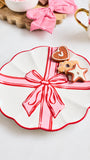 A scalloped white ceramic plate decorated with a red border and a pink gift box design with a red ribbon and bow, styled with cookies on a light tabletop.