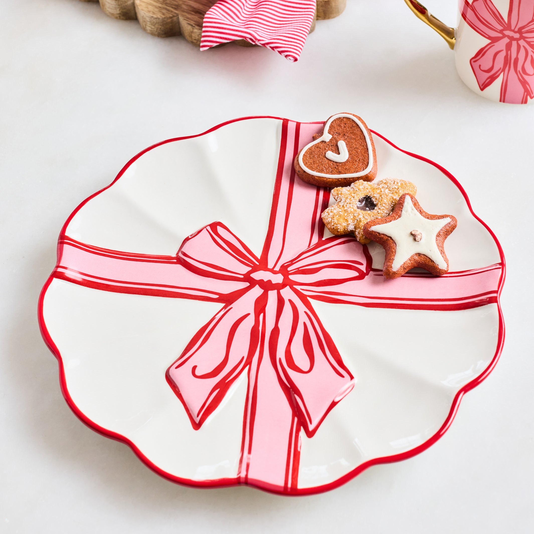 A scalloped white ceramic plate decorated with a red border and a pink gift box design with a red ribbon and bow, styled with cookies on a light tabletop.