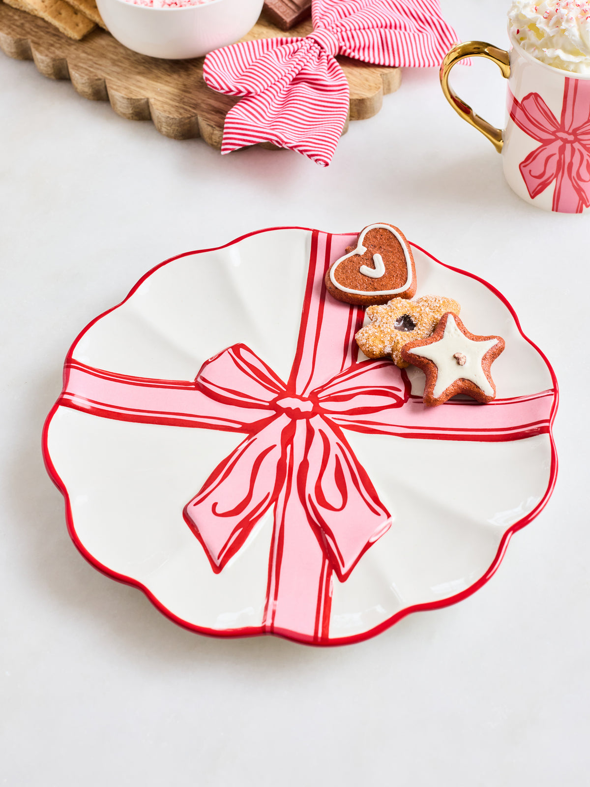 A scalloped white ceramic plate decorated with a red border and a pink gift box design with a red ribbon and bow, styled with cookies on a light tabletop.