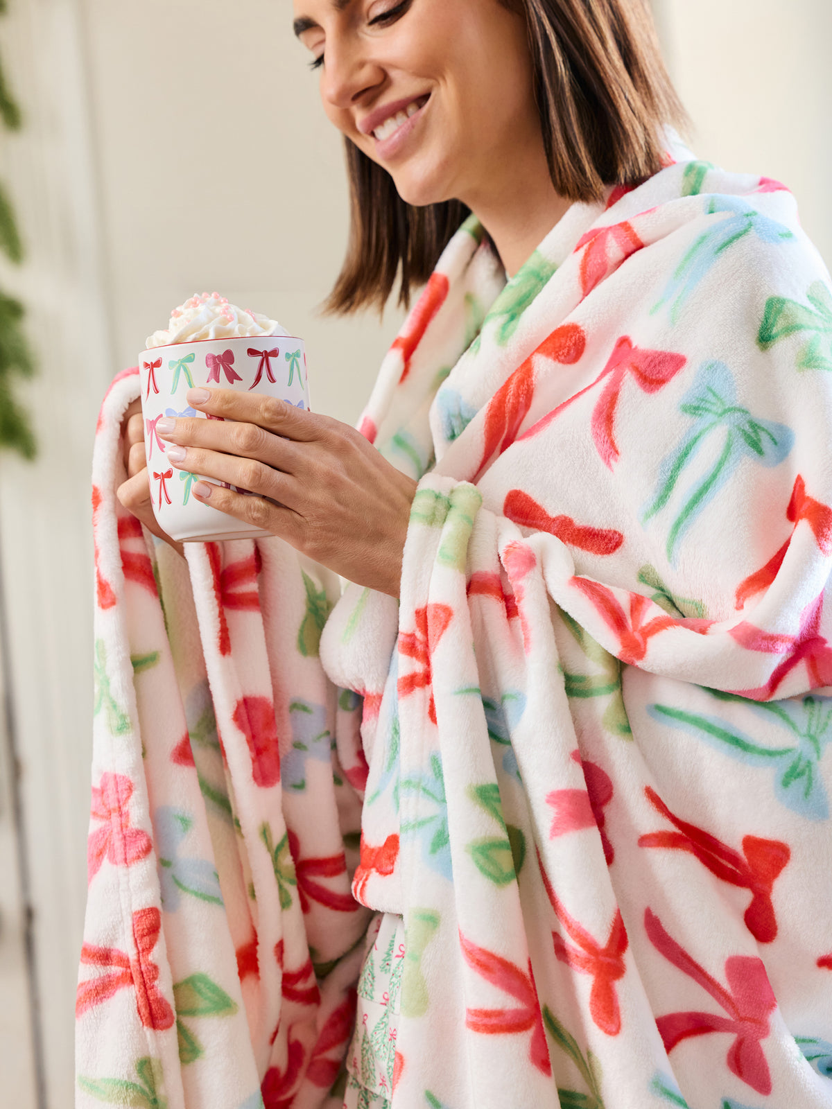 A woman wrapped in a soft fleece blanket with a colorful dragonfly pattern, holding a mug topped with whipped cream and hot chocolate.