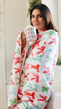 A woman wrapped in a white fleece robe with a colorful Christmas pattern of red, green, and pink ornaments, holding a mug in a bright bathroom setting.