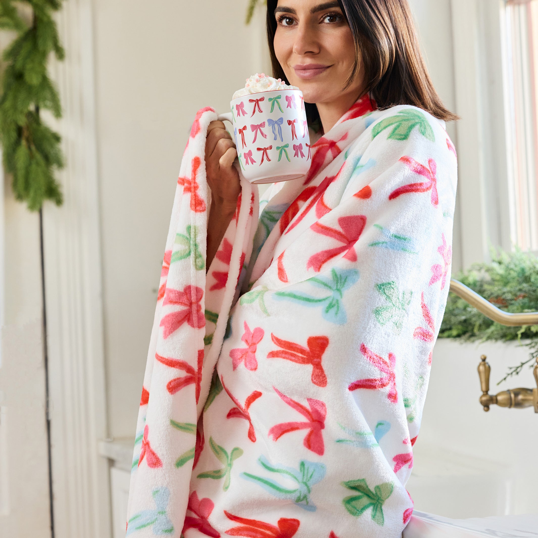 A woman wrapped in a white fleece robe with a colorful Christmas pattern of red, green, and pink ornaments, holding a mug in a bright bathroom setting.