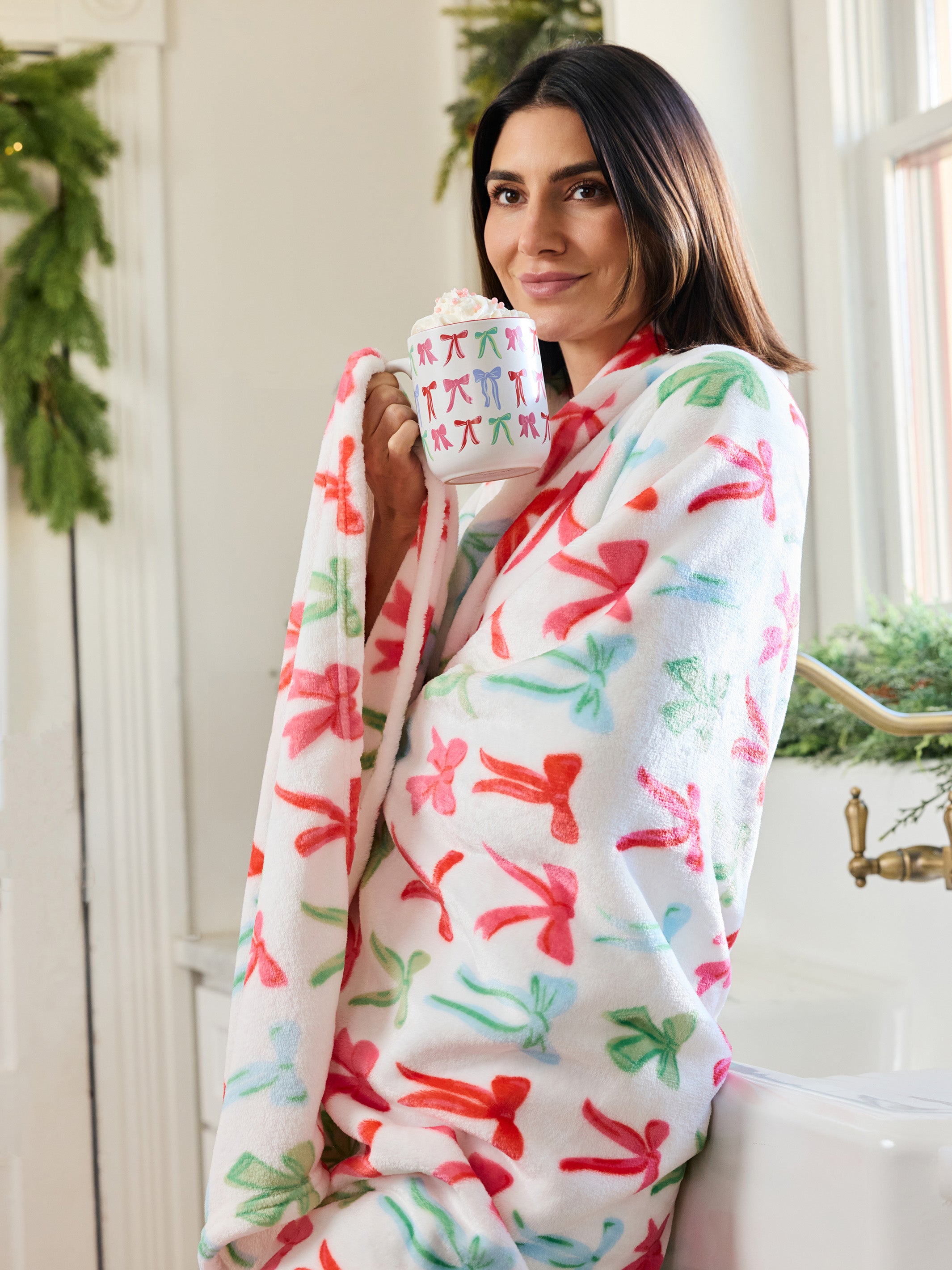 A woman wrapped in a white fleece robe with a colorful Christmas pattern of red, green, and pink ornaments, holding a mug in a bright bathroom setting.