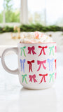 A white ceramic mug filled with whipped cream and a pink drink, decorated with a repeating pattern of colorful bows in red, green, blue, and pink, set on a light marble countertop with a blurred kitchen background.
