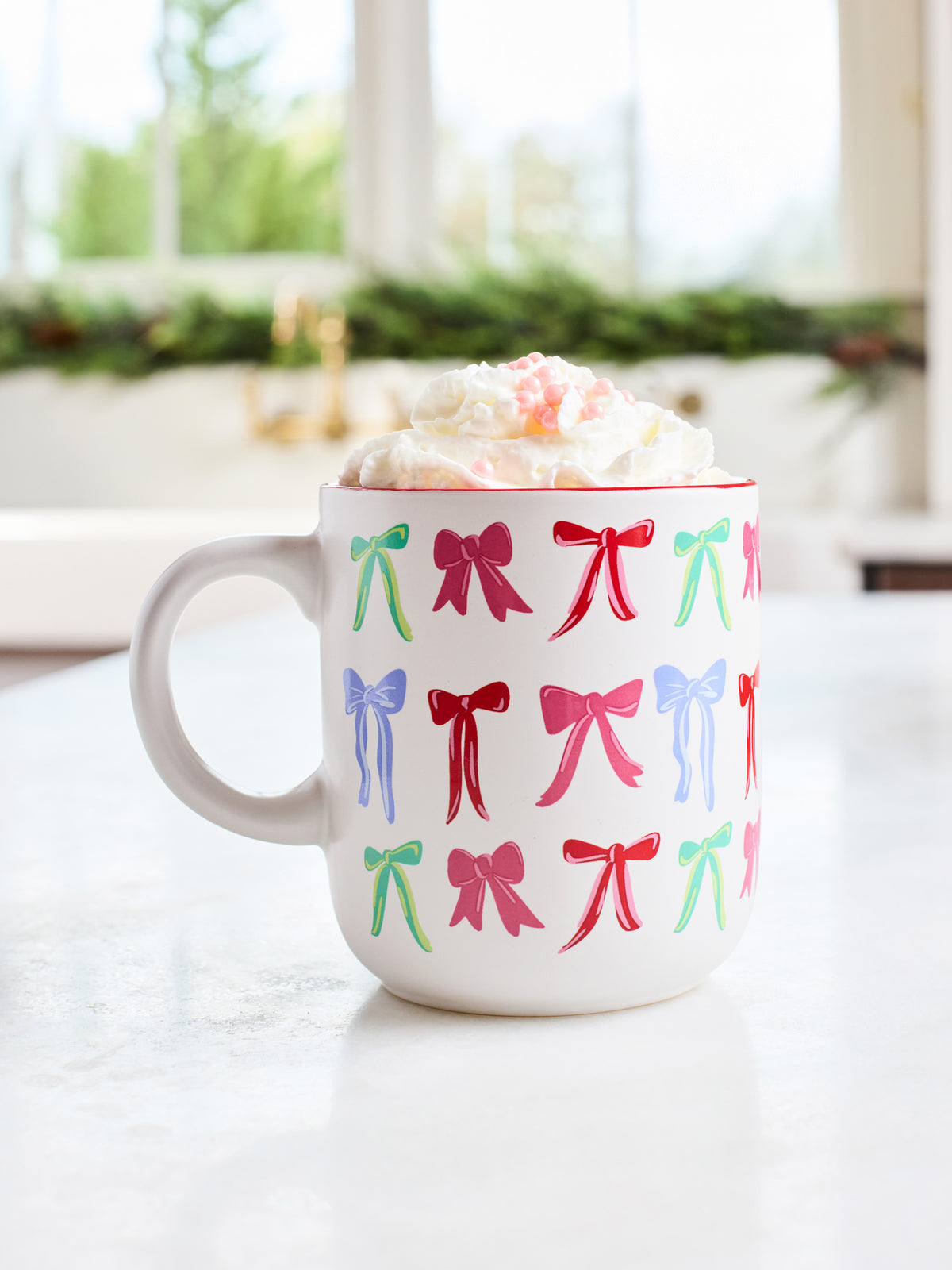 A white ceramic mug filled with whipped cream and a pink drink, decorated with a repeating pattern of colorful bows in red, green, blue, and pink, set on a light marble countertop with a blurred kitchen background.