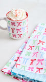 A white ceramic mug filled with whipped cream and candy, placed next to a stack of folded fabric napkins or tea towels printed with colorful bow motifs on a white background.