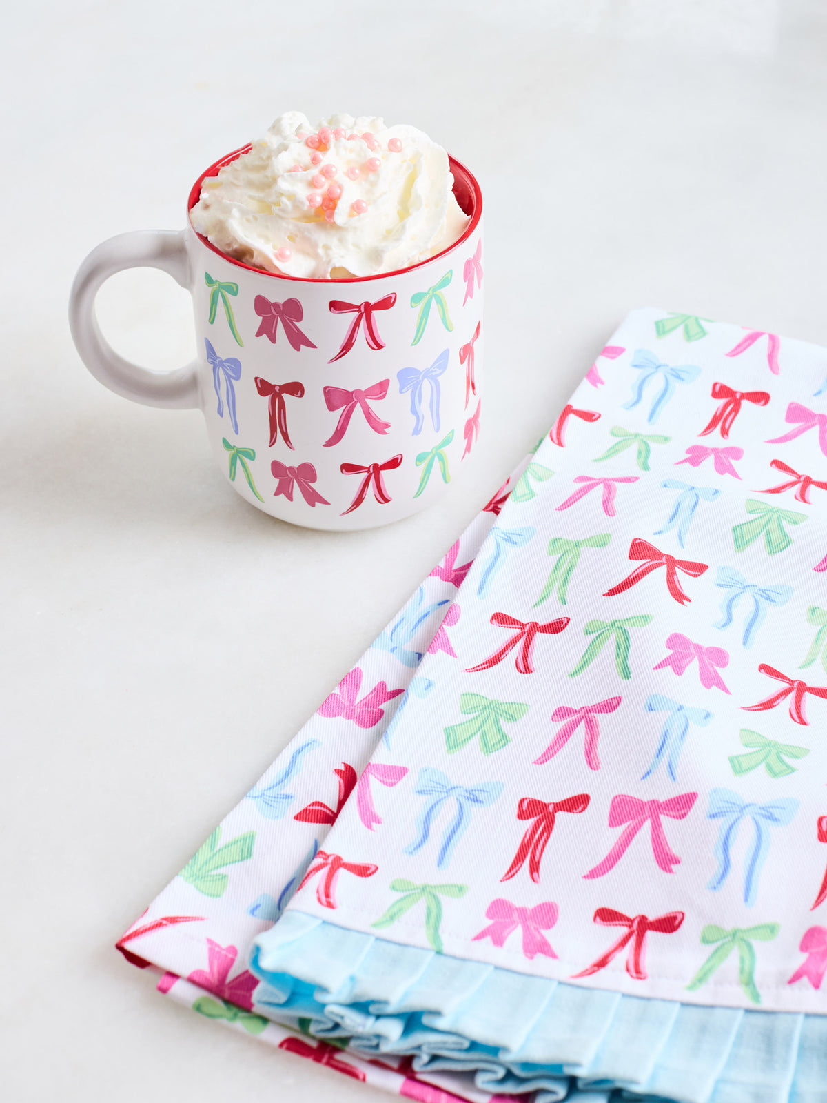 A white ceramic mug filled with whipped cream and candy, placed next to a stack of folded fabric napkins or tea towels printed with colorful bow motifs on a white background.