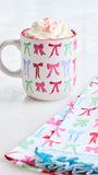 A white ceramic mug with a red rim, decorated with a repeating pattern of colorful bows in pink, green, and blue. The mug is filled with whipped cream and pink sprinkles, set on a light surface next to folded napkins with the same bow pattern.