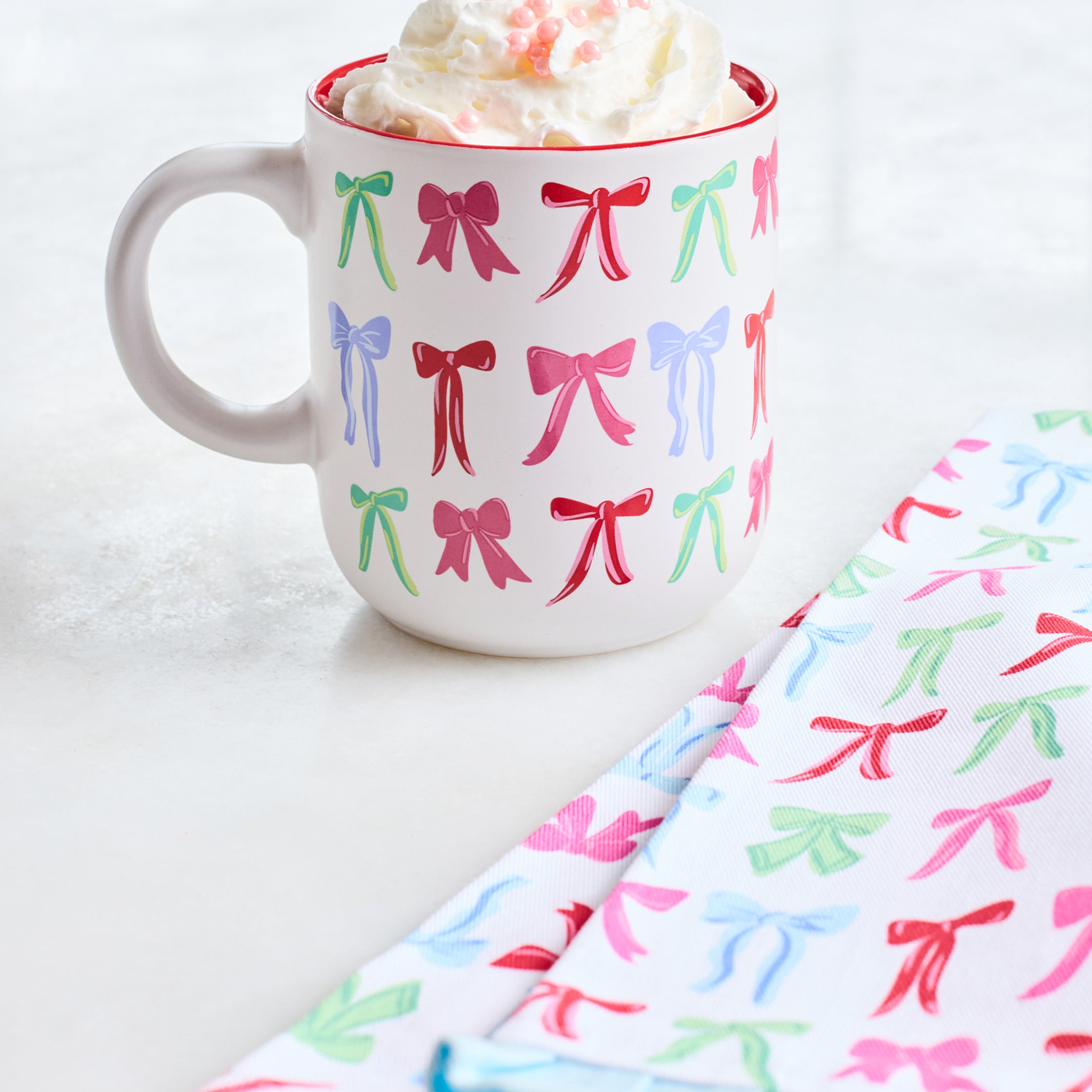 A white ceramic mug with a red rim, decorated with a repeating pattern of colorful bows in pink, green, and blue. The mug is filled with whipped cream and pink sprinkles, set on a light surface next to folded napkins with the same bow pattern.
