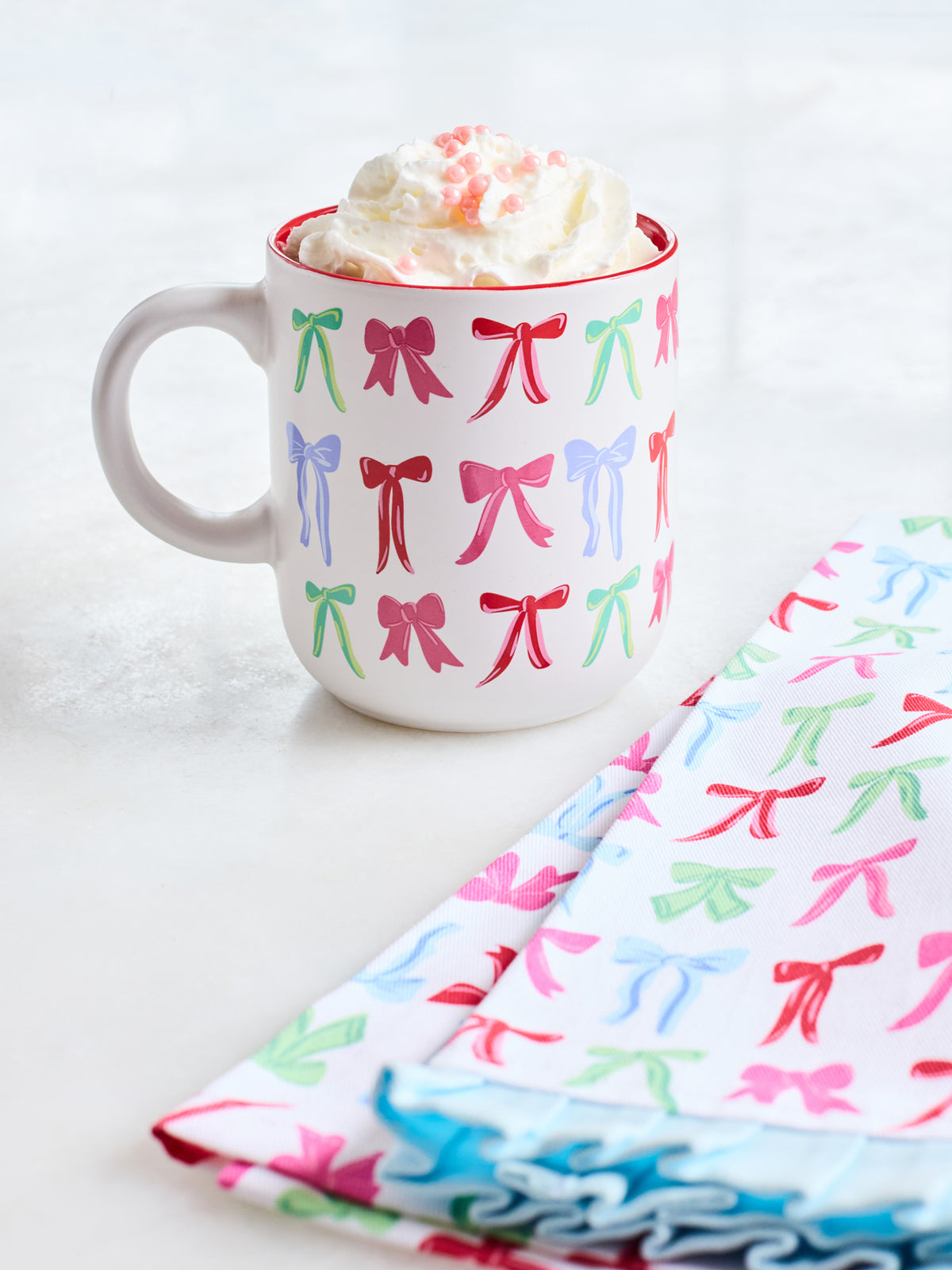 A white ceramic mug with a red rim, decorated with a repeating pattern of colorful bows in pink, green, and blue. The mug is filled with whipped cream and pink sprinkles, set on a light surface next to folded napkins with the same bow pattern.