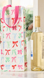 A white wine gift bag with pink handles and a repeating pattern of colorful bow illustrations, standing next to a bottle of white wine on a light surface.