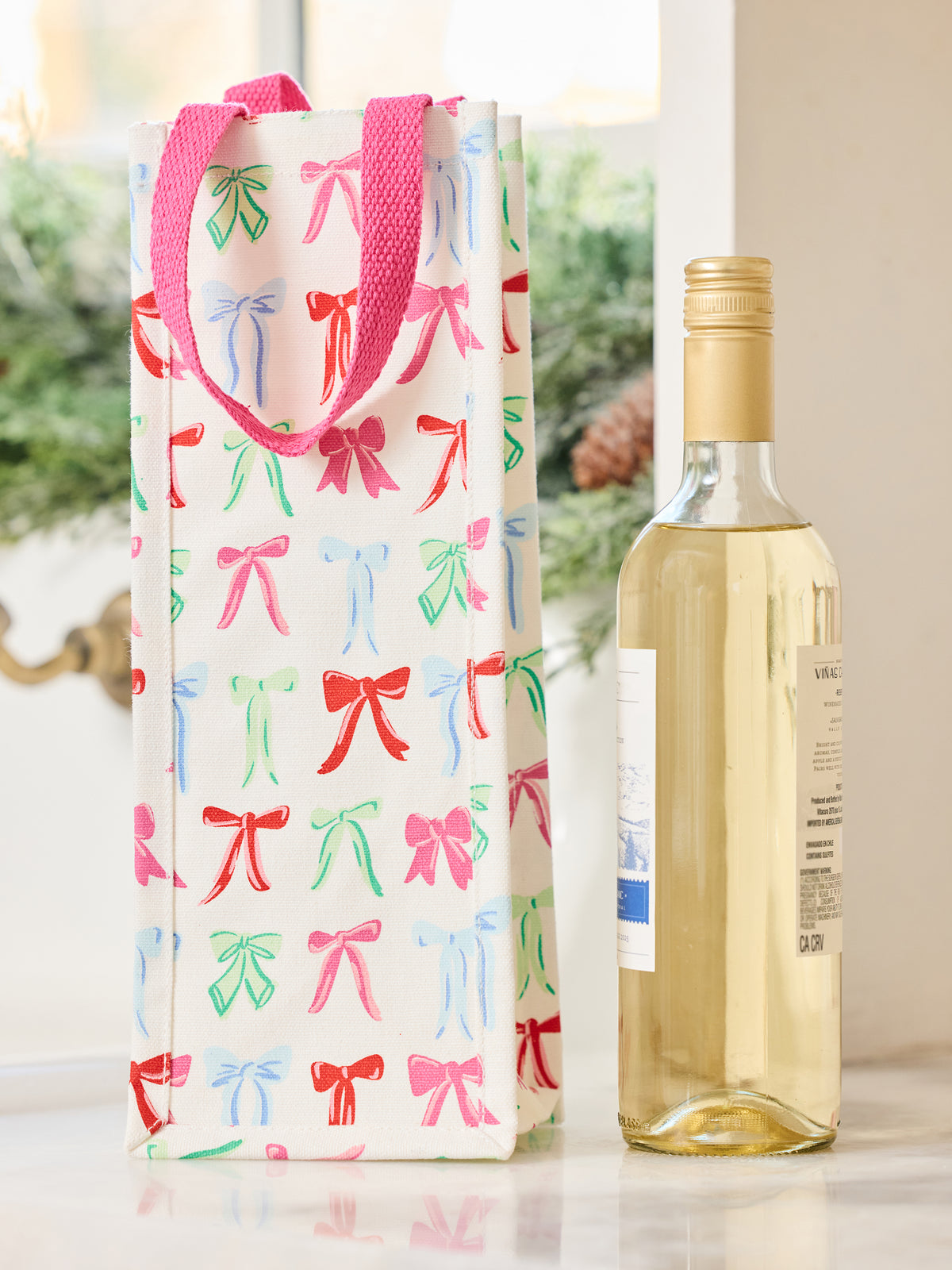 A white wine gift bag with pink handles and a repeating pattern of colorful bow illustrations, standing next to a bottle of white wine on a light surface.