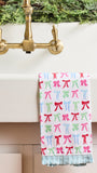 A white kitchen towel with a colorful pattern of pink, red, and green bows, hanging over the edge of a white sink with a brass faucet.