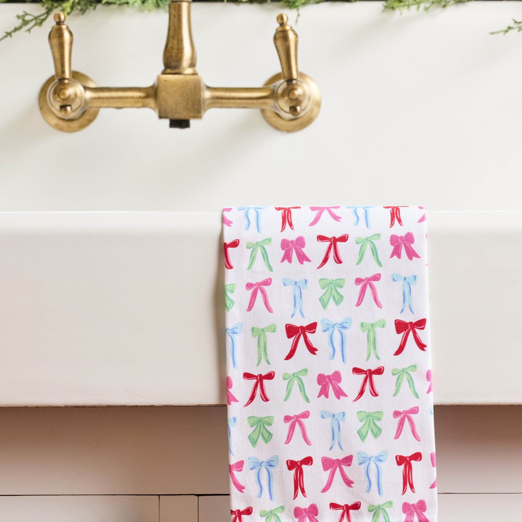 A white kitchen towel with a colorful pattern of pink, red, and green bows, hanging over the edge of a white sink with a brass faucet.