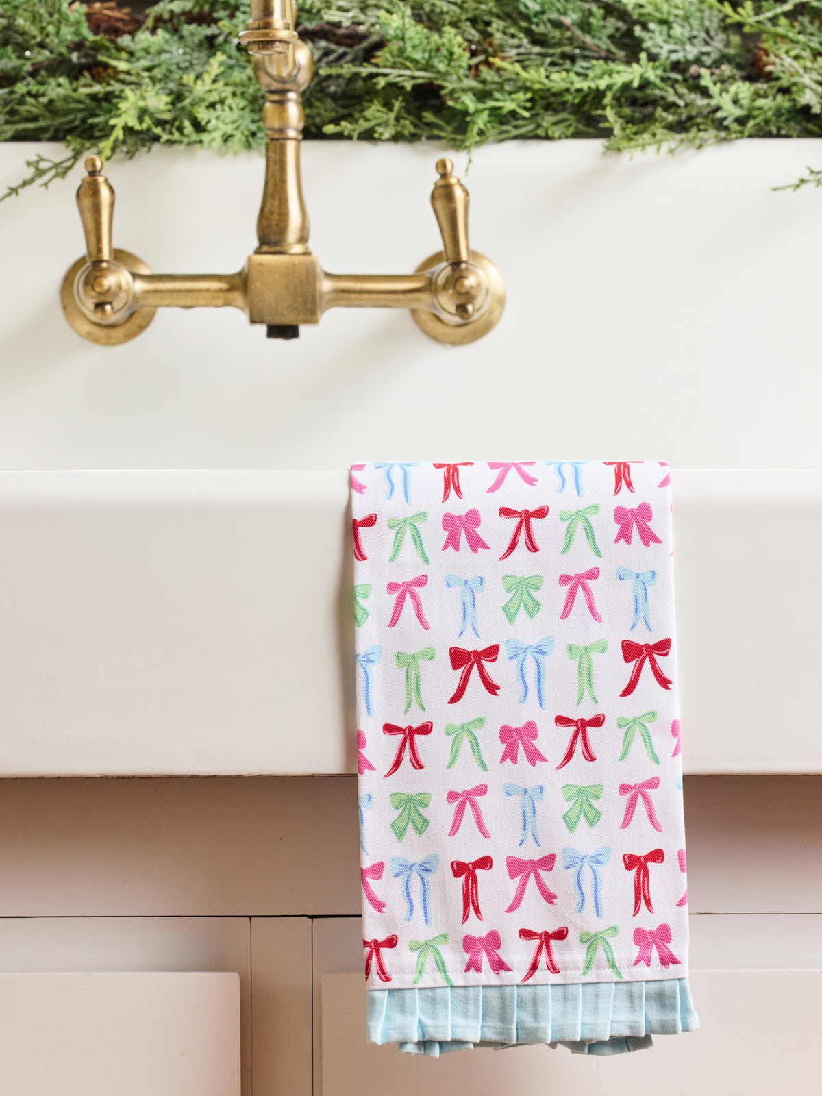 A white kitchen towel with a colorful pattern of pink, red, and green bows, hanging over the edge of a white sink with a brass faucet.
