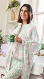 A woman stands in a bright kitchen wearing a white pajama set with a green and pink Christmas tree and bow pattern, holding a green mug.