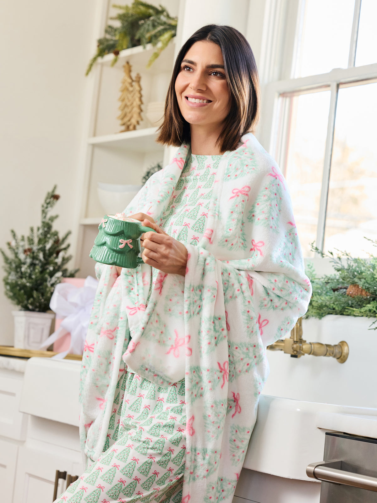 A woman stands in a bright kitchen wearing a white pajama set with a green and pink Christmas tree and bow pattern, holding a green mug.