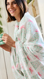 A woman wrapped in a soft white fleece blanket with a festive green and pink Christmas tree pattern, holding a green mug in a bright indoor setting.