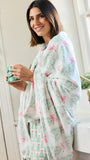 A woman wrapped in a soft white robe with a green and pink Christmas tree and bow pattern, holding a green mug in a bright bathroom setting.