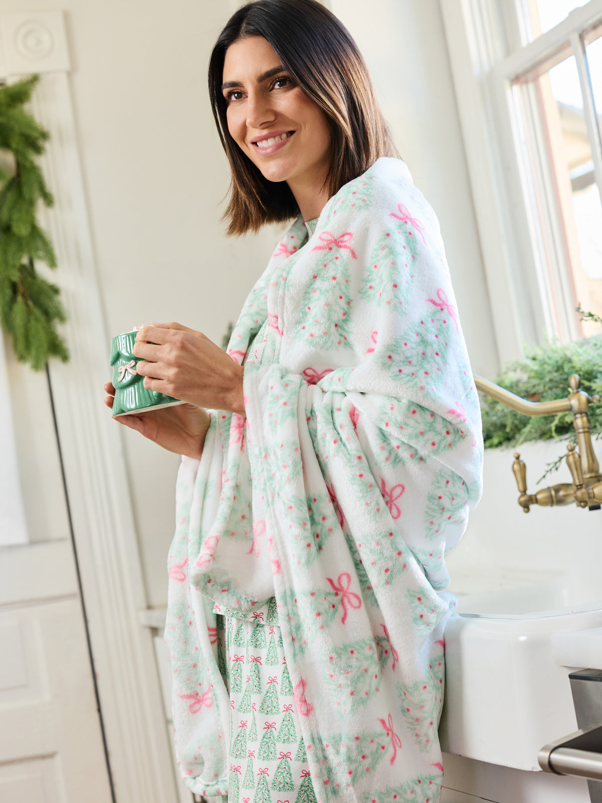 A woman wrapped in a soft white robe with a green and pink Christmas tree and bow pattern, holding a green mug in a bright bathroom setting.