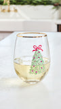A clear stemless wine glass with a gold rim, filled with white wine and decorated with a green Christmas tree illustration topped with a red bow, set on a light countertop in a bright kitchen.